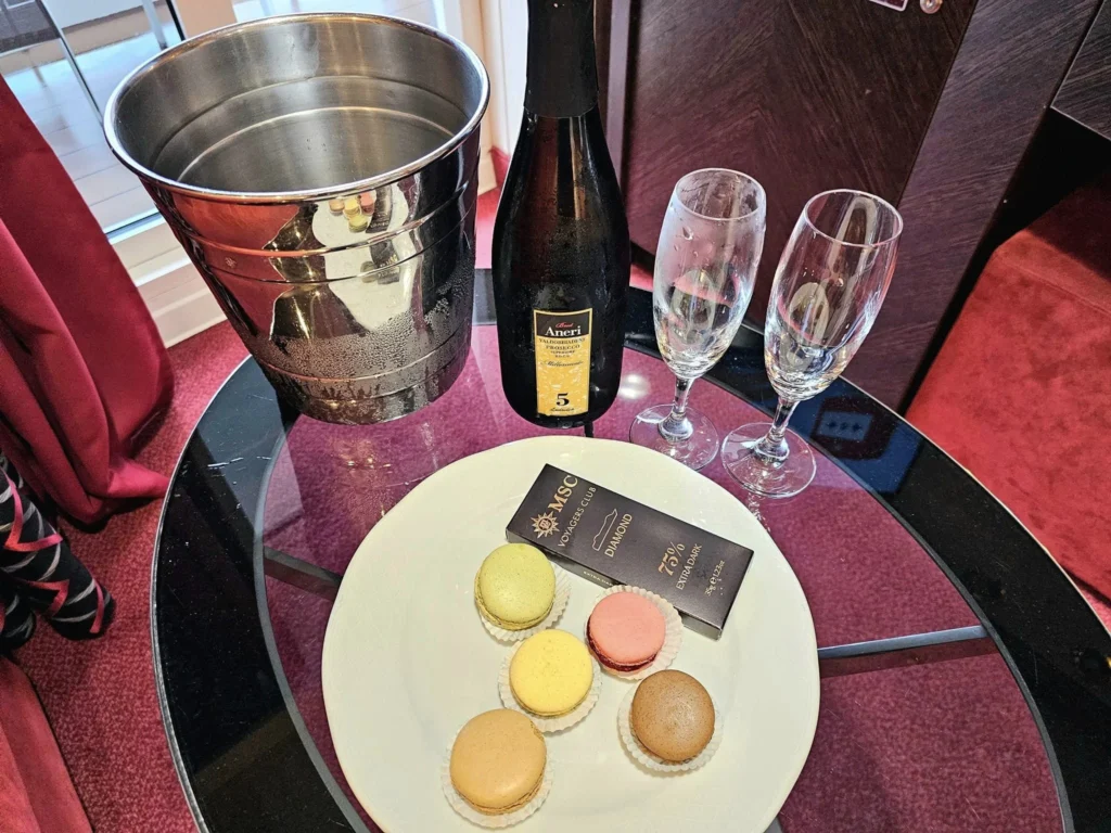 Cruising with Danny and Peter, MSC Cruises, MSC Preziosa, Rotterdam, The Netherlands, Birthday Cruise | The majestic MSC Preziosa cruise ship docked at Rotterdam’s Cruise Terminal on Wilhelminakade, its sleek white exterior gleaming against the modern city skyline with the iconic Erasmus Bridge in the background. Passengers board for a Northern Europe mini-cruise, celebrating a birthday with luxurious touches like a chilled bottle of Spumante and macarons in a stylish cabin. The vibrant onboard atmosphere features a gourmet dining experience at Butcher’s Cut Chef’s Table with a surprise birthday cake, thrilling entertainment at the Platinum Theater’s Rock Nation show with singers and dancers, and a lively Silent Disco in the Safari Lounge. The scene captures the excitement of embarkation day, with scenic views of Rotterdam’s waterfront, onboard shopping for MSC souvenirs, and the promise of an unforgettable luxury cruise vacation.