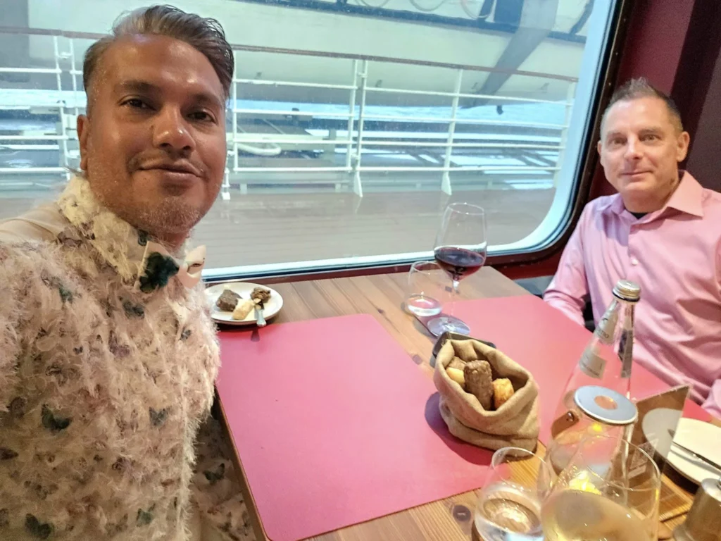 Cruising with Danny and Peter, MSC Cruises, MSC Preziosa, Rotterdam, The Netherlands, Birthday Cruise | The majestic MSC Preziosa cruise ship docked at Rotterdam’s Cruise Terminal on Wilhelminakade, its sleek white exterior gleaming against the modern city skyline with the iconic Erasmus Bridge in the background. Passengers board for a Northern Europe mini-cruise, celebrating a birthday with luxurious touches like a chilled bottle of Spumante and macarons in a stylish cabin. The vibrant onboard atmosphere features a gourmet dining experience at Butcher’s Cut Chef’s Table with a surprise birthday cake, thrilling entertainment at the Platinum Theater’s Rock Nation show with singers and dancers, and a lively Silent Disco in the Safari Lounge. The scene captures the excitement of embarkation day, with scenic views of Rotterdam’s waterfront, onboard shopping for MSC souvenirs, and the promise of an unforgettable luxury cruise vacation.