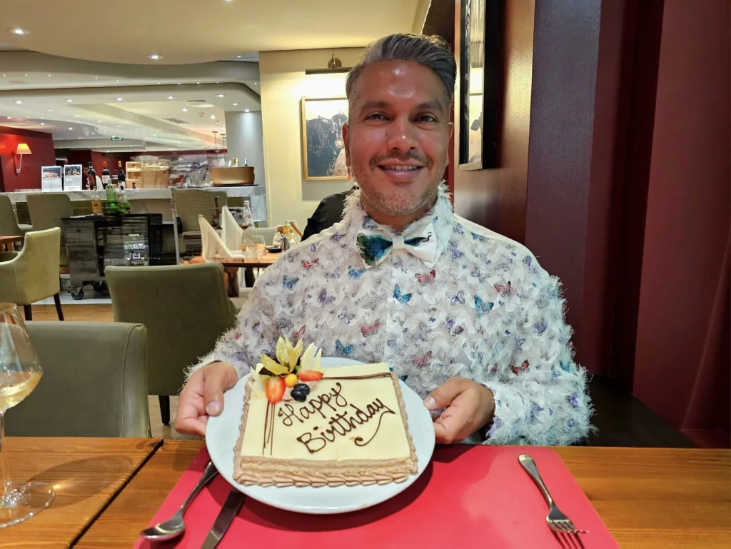 Cruising with Danny and Peter, MSC Cruises, MSC Preziosa, Rotterdam, The Netherlands, Birthday Cruise | The majestic MSC Preziosa cruise ship docked at Rotterdam’s Cruise Terminal on Wilhelminakade, its sleek white exterior gleaming against the modern city skyline with the iconic Erasmus Bridge in the background. Passengers board for a Northern Europe mini-cruise, celebrating a birthday with luxurious touches like a chilled bottle of Spumante and macarons in a stylish cabin. The vibrant onboard atmosphere features a gourmet dining experience at Butcher’s Cut Chef’s Table with a surprise birthday cake, thrilling entertainment at the Platinum Theater’s Rock Nation show with singers and dancers, and a lively Silent Disco in the Safari Lounge. The scene captures the excitement of embarkation day, with scenic views of Rotterdam’s waterfront, onboard shopping for MSC souvenirs, and the promise of an unforgettable luxury cruise vacation.