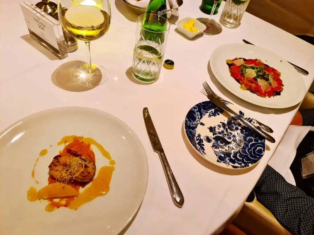 Cruising with Danny and Peter, Holland America Line, Koningsdam, Pacific Ocean, Sea Day | A breathtaking view of the ocean during a Relaxing Cruise Day at Sea Adventure, showcasing scenic ocean views and the cruise ship gliding over the waves. Guests enjoy onboard activities, sipping cocktails by the poolside, and indulging in a gourmet cruise ship dining experience in the elegant dining room, surrounded by a vibrant atmosphere and happy passengers. The sun sets in the background, adding warmth to the leisure at sea.