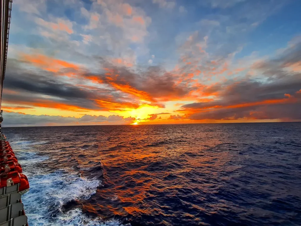 Cruising with Danny and Peter, Holland America Line, Koningsdam, Pacific Ocean, Sea Day | A stunning sunrise over the ocean during a Hawaiian cruise, showcasing the vibrant colors of the sky, a luxury cruise ship on calm waters, guests enjoying marine biology lectures, participating in ukulele lessons, and savoring delicious culinary experiences, embodying relaxation and adventure at sea in the beautiful Hawaiian Islands.