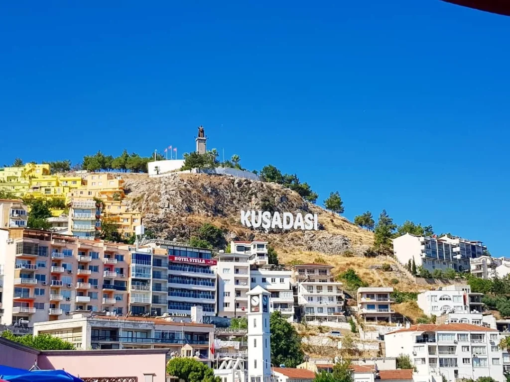 Cruising with Danny and Peter, Holland America Line, Koningsdam, Kusadasi, Ephesus, Turkey | A picturesque view of Kuşadası Beach showcasing soft golden sands and crystal-clear Aegean Sea waters, vibrant local markets filled with traditional Turkish crafts, and visitors savoring authentic Turkish cuisine at a charming café, highlighting the cultural exploration and seaside relaxation of a perfect Kuşadası Beach Day on the Turkish Riviera.