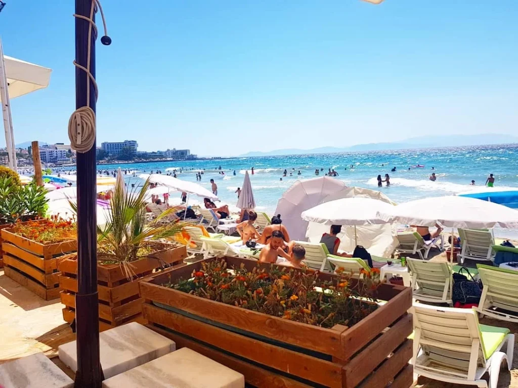 Cruising with Danny and Peter, Holland America Line, Koningsdam, Kusadasi, Ephesus, Turkey | A picturesque view of Kuşadası Beach showcasing soft golden sands and crystal-clear Aegean Sea waters, vibrant local markets filled with traditional Turkish crafts, and visitors savoring authentic Turkish cuisine at a charming café, highlighting the cultural exploration and seaside relaxation of a perfect Kuşadası Beach Day on the Turkish Riviera.