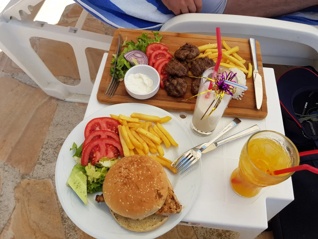 Cruising with Danny and Peter, Holland America Line, Koningsdam, Kusadasi, Ephesus, Turkey | A picturesque view of Kuşadası Beach showcasing soft golden sands and crystal-clear Aegean Sea waters, vibrant local markets filled with traditional Turkish crafts, and visitors savoring authentic Turkish cuisine at a charming café, highlighting the cultural exploration and seaside relaxation of a perfect Kuşadası Beach Day on the Turkish Riviera.