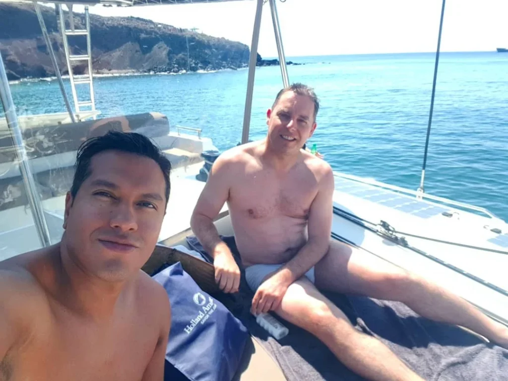 Cruising with Danny and Peter, Holland America Line, Koningsdam, Thira, Santorini, Greece | Luxury catamaran cruising through Santorini’s volcanic caldera, showcasing whitewashed buildings perched on cliffs, a secluded cove with warm mineral-rich waters, and guests enjoying a stunning sunset while savoring local delicacies prepared by the onboard chef during an exclusive sailing experience.