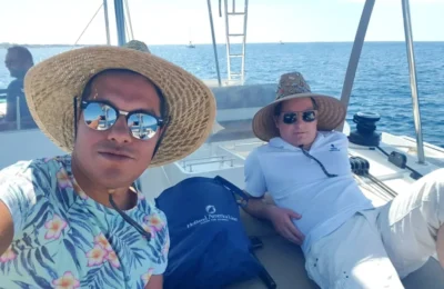 Cruising with Danny and Peter, Holland America Line, Koningsdam, Thira, Santorini, Greece | Luxury catamaran cruising through Santorini’s volcanic caldera, showcasing whitewashed buildings perched on cliffs, a secluded cove with warm mineral-rich waters, and guests enjoying a stunning sunset while savoring local delicacies prepared by the onboard chef during an exclusive sailing experience.