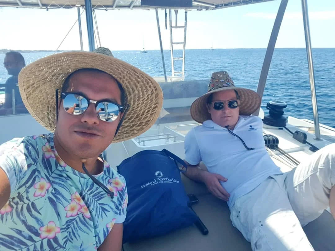 Cruising with Danny and Peter, Holland America Line, Koningsdam, Thira, Santorini, Greece | Luxury catamaran cruising through Santorini’s volcanic caldera, showcasing whitewashed buildings perched on cliffs, a secluded cove with warm mineral-rich waters, and guests enjoying a stunning sunset while savoring local delicacies prepared by the onboard chef during an exclusive sailing experience.