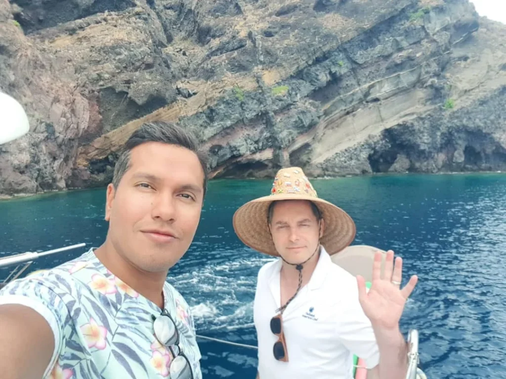 Cruising with Danny and Peter, Holland America Line, Koningsdam, Thira, Santorini, Greece | Luxury catamaran cruising through Santorini’s volcanic caldera, showcasing whitewashed buildings perched on cliffs, a secluded cove with warm mineral-rich waters, and guests enjoying a stunning sunset while savoring local delicacies prepared by the onboard chef during an exclusive sailing experience.