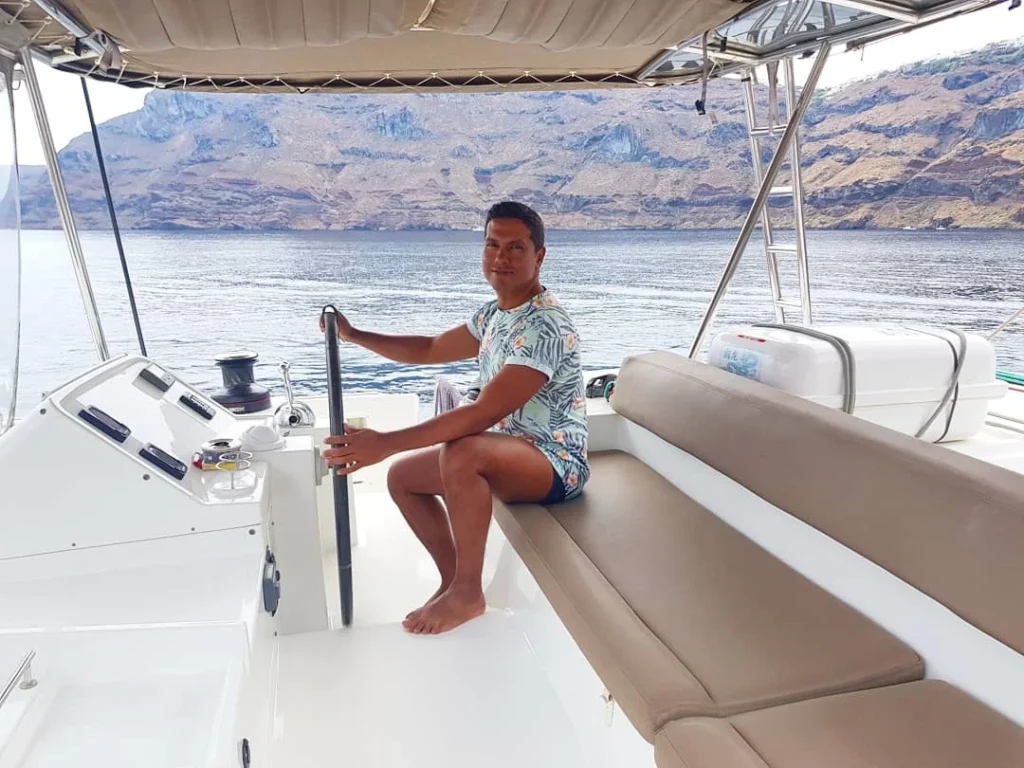 Cruising with Danny and Peter, Holland America Line, Koningsdam, Thira, Santorini, Greece | Luxury catamaran cruising through Santorini’s volcanic caldera, showcasing whitewashed buildings perched on cliffs, a secluded cove with warm mineral-rich waters, and guests enjoying a stunning sunset while savoring local delicacies prepared by the onboard chef during an exclusive sailing experience.