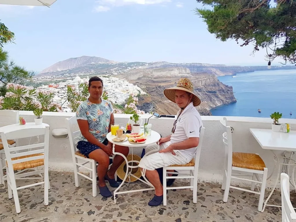 Cruising with Danny and Peter, Holland America Line, Koningsdam, Thira, Santorini, Greece | Luxury catamaran cruising through Santorini’s volcanic caldera, showcasing whitewashed buildings perched on cliffs, a secluded cove with warm mineral-rich waters, and guests enjoying a stunning sunset while savoring local delicacies prepared by the onboard chef during an exclusive sailing experience.