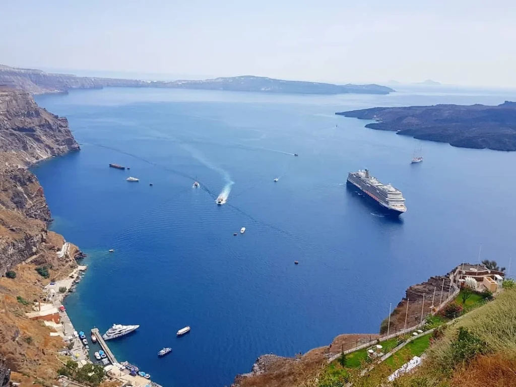 Cruising with Danny and Peter, Holland America Line, Koningsdam, Thira, Santorini, Greece | Luxury catamaran cruising through Santorini’s volcanic caldera, showcasing whitewashed buildings perched on cliffs, a secluded cove with warm mineral-rich waters, and guests enjoying a stunning sunset while savoring local delicacies prepared by the onboard chef during an exclusive sailing experience.