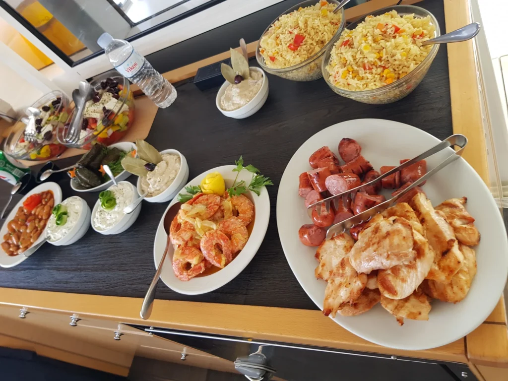 Cruising with Danny and Peter, Holland America Line, Koningsdam, Thira, Santorini, Greece | Luxury catamaran cruising through Santorini’s volcanic caldera, showcasing whitewashed buildings perched on cliffs, a secluded cove with warm mineral-rich waters, and guests enjoying a stunning sunset while savoring local delicacies prepared by the onboard chef during an exclusive sailing experience.