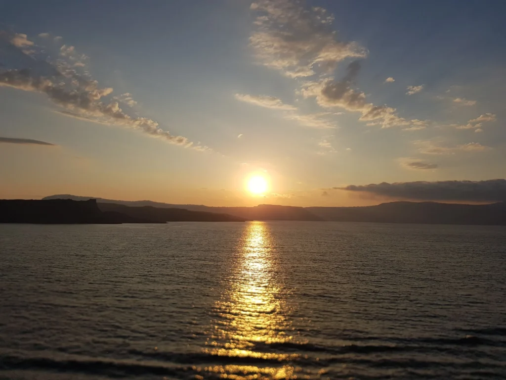 Cruising with Danny and Peter, Holland America Line, Koningsdam, Thira, Santorini, Greece | Luxury catamaran cruising through Santorini’s volcanic caldera, showcasing whitewashed buildings perched on cliffs, a secluded cove with warm mineral-rich waters, and guests enjoying a stunning sunset while savoring local delicacies prepared by the onboard chef during an exclusive sailing experience.