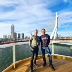 Cruising with Danny and Peter, Holland America Line, Rotterdam, Rotterdam, The Netherlands | A stunning view of the M.S. Rotterdam as it sails away from Rotterdam, celebrating Holland America 150th Anniversary with historic ships in the background. The sunset casts a golden glow on the luxury ocean liner, capturing the excitement of our transatlantic voyage from Rotterdam to New York. A special anniversary menu and Holland America memorabilia highlight this remarkable journey.