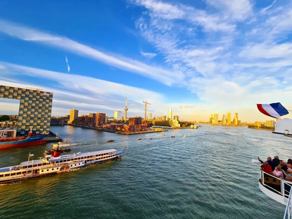 Cruising with Danny and Peter, Holland America Line, Rotterdam, Rotterdam, The Netherlands | A stunning view of the M.S. Rotterdam as it sails away from Rotterdam, celebrating Holland America 150th Anniversary with historic ships in the background. The sunset casts a golden glow on the luxury ocean liner, capturing the excitement of our transatlantic voyage from Rotterdam to New York. A special anniversary menu and Holland America memorabilia highlight this remarkable journey.