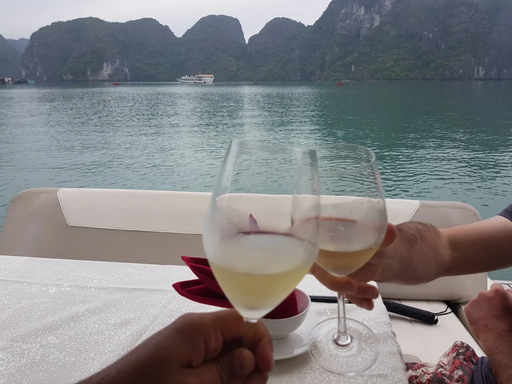 Cruising with Danny and Peter, Holland America Line, Westerdam, Halong Bay, Vietnam | A stunning view of the Hạ Long Bay Yacht cruising through emerald waters, surrounded by majestic limestone karsts. Guests enjoy Vietnamese seafood dining on the deck while kayaking in Hạ Long Bay. The private yacht tour showcases the beauty of this UNESCO World Heritage site.