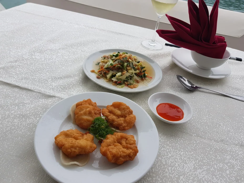 Cruising with Danny and Peter, Holland America Line, Westerdam, Halong Bay, Vietnam | A stunning view of the Hạ Long Bay Yacht cruising through emerald waters, surrounded by majestic limestone karsts. Guests enjoy Vietnamese seafood dining on the deck while kayaking in Hạ Long Bay. The private yacht tour showcases the beauty of this UNESCO World Heritage site.