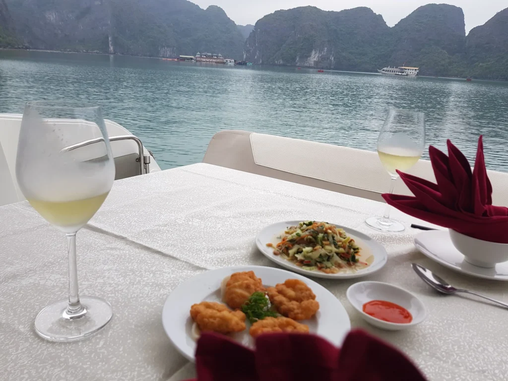 Cruising with Danny and Peter, Holland America Line, Westerdam, Halong Bay, Vietnam | A stunning view of the Hạ Long Bay Yacht cruising through emerald waters, surrounded by majestic limestone karsts. Guests enjoy Vietnamese seafood dining on the deck while kayaking in Hạ Long Bay. The private yacht tour showcases the beauty of this UNESCO World Heritage site.