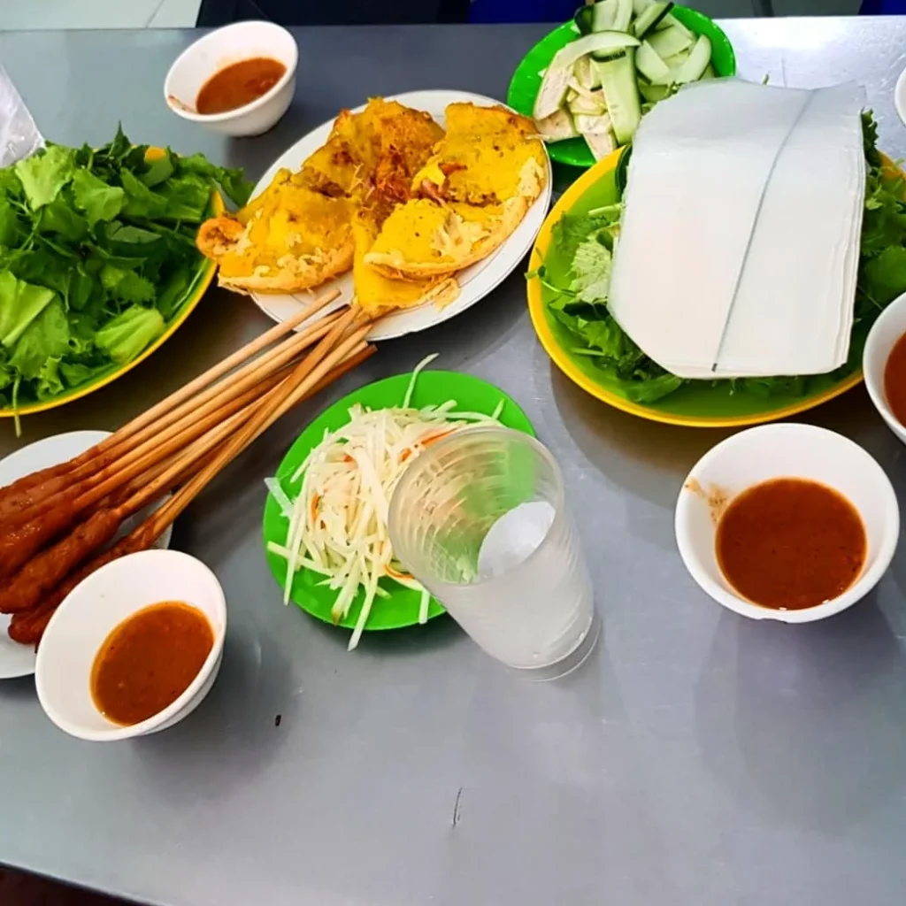 Cruising with Danny and Peter, Holland America Line, Westerdam, Da Nang, Hue, Vietnam | A vibrant street scene in Đà Nẵng showcasing a bustling market filled with fresh produce and aromatic spices, capturing the essence of Vietnamese cuisine. Motorbikes are parked nearby, symbolizing the thrilling motorbike street food tour. An enticing plate of bánh mì and bowls of pho are featured prominently, representing the local street eats enjoyed during this culinary exploration.