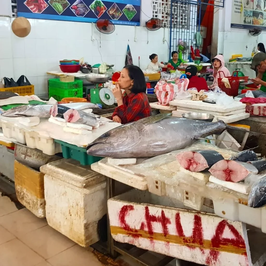 Cruising with Danny and Peter, Holland America Line, Westerdam, Da Nang, Hue, Vietnam | A vibrant street scene in Đà Nẵng showcasing a bustling market filled with fresh produce and aromatic spices, capturing the essence of Vietnamese cuisine. Motorbikes are parked nearby, symbolizing the thrilling motorbike street food tour. An enticing plate of bánh mì and bowls of pho are featured prominently, representing the local street eats enjoyed during this culinary exploration.