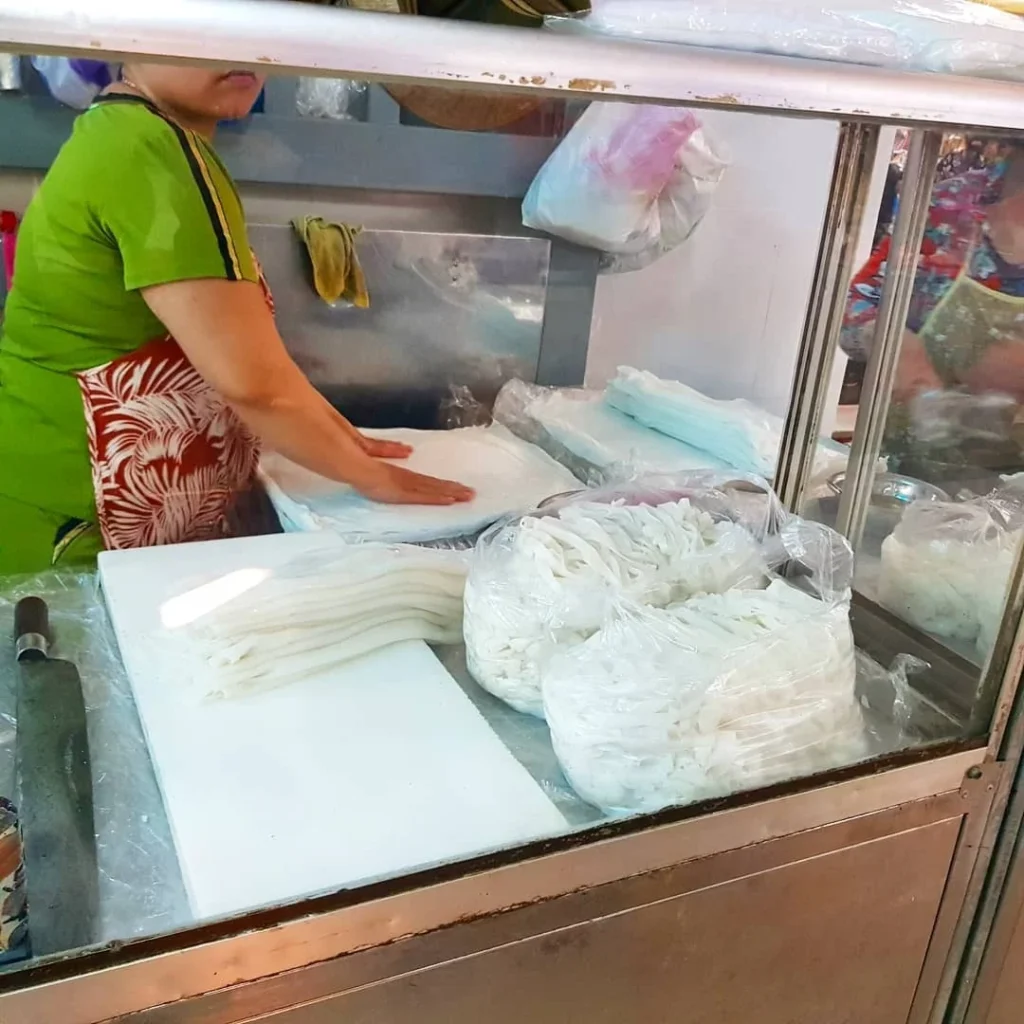 Cruising with Danny and Peter, Holland America Line, Westerdam, Da Nang, Hue, Vietnam | A vibrant street scene in Đà Nẵng showcasing a bustling market filled with fresh produce and aromatic spices, capturing the essence of Vietnamese cuisine. Motorbikes are parked nearby, symbolizing the thrilling motorbike street food tour. An enticing plate of bánh mì and bowls of pho are featured prominently, representing the local street eats enjoyed during this culinary exploration.