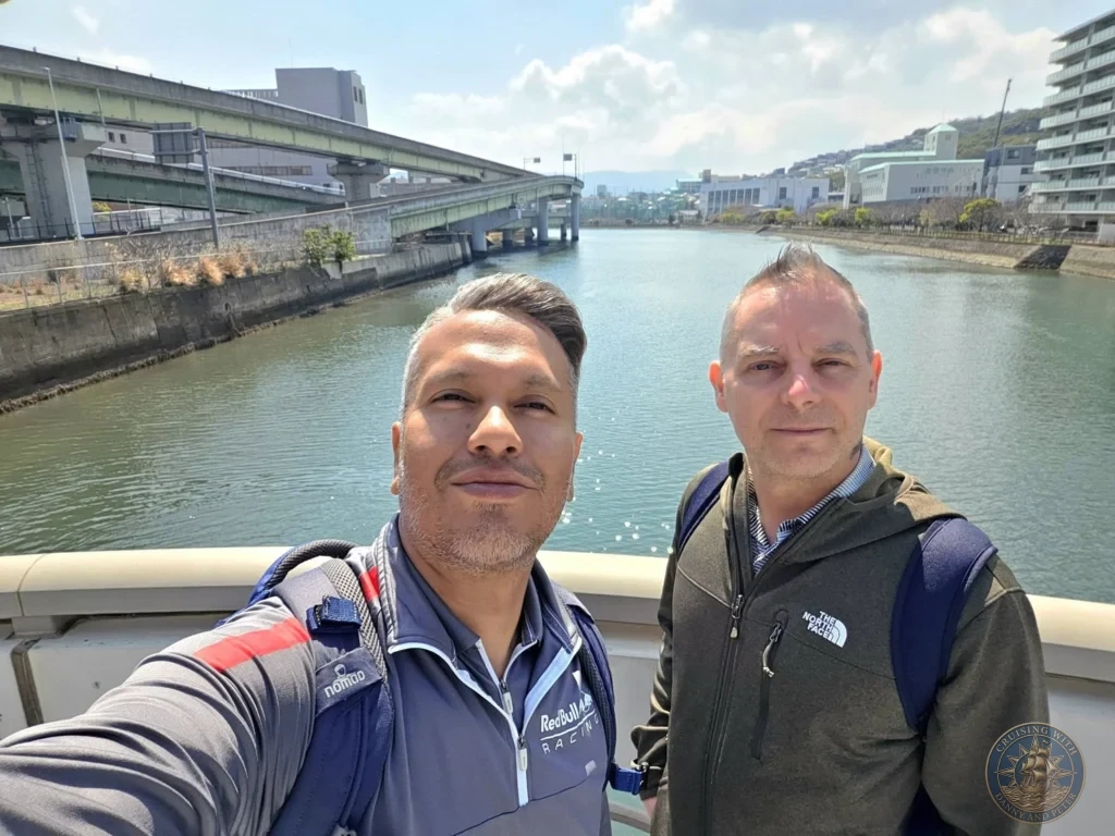Selfie of Danny and Peter at Mirai Nagasaki Coco Walk waterfront in Nagasaki Japan cruise port guide. Perfect spot for things to do in Nagasaki from cruise ship and one day in Nagasaki Japan itinerary. Westerdam passengers enjoying the sunny day with city river, bridges, and Ferris wheel area in background by Danny and Peter.
