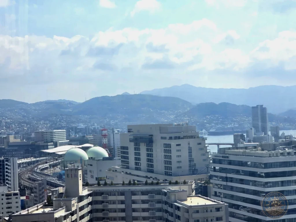 Panoramic view from the Nagasaki Ferris wheel showing the city skyline, harbor with the Westrdam, modern buildings, train tracks, and green mountains in the distance — featured in Nagasaki Japan cruise port guide. Ideal for things to do in Nagasaki from cruise ship and one day in Nagasaki Japan itinerary from the Westerdam docking location by Danny and Peter.