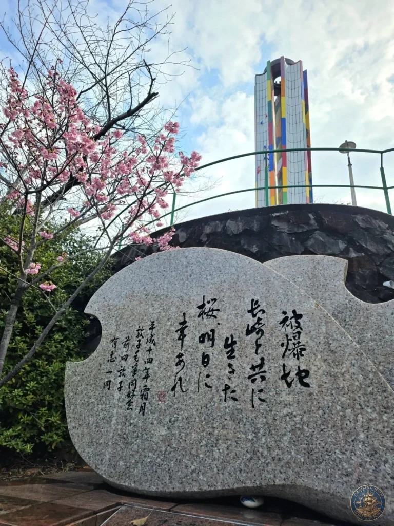 Large inscribed stone monument at the Hypocenter Park in Nagasaki, Japan, with Japanese calligraphy, blooming cherry blossom tree in the foreground, and the colorful Peace Tower in the background. Featured in Nagasaki Japan cruise port guide, things to do in Nagasaki from cruise ship, and one day in Nagasaki Japan itinerary from the Westerdam by Danny and Peter.