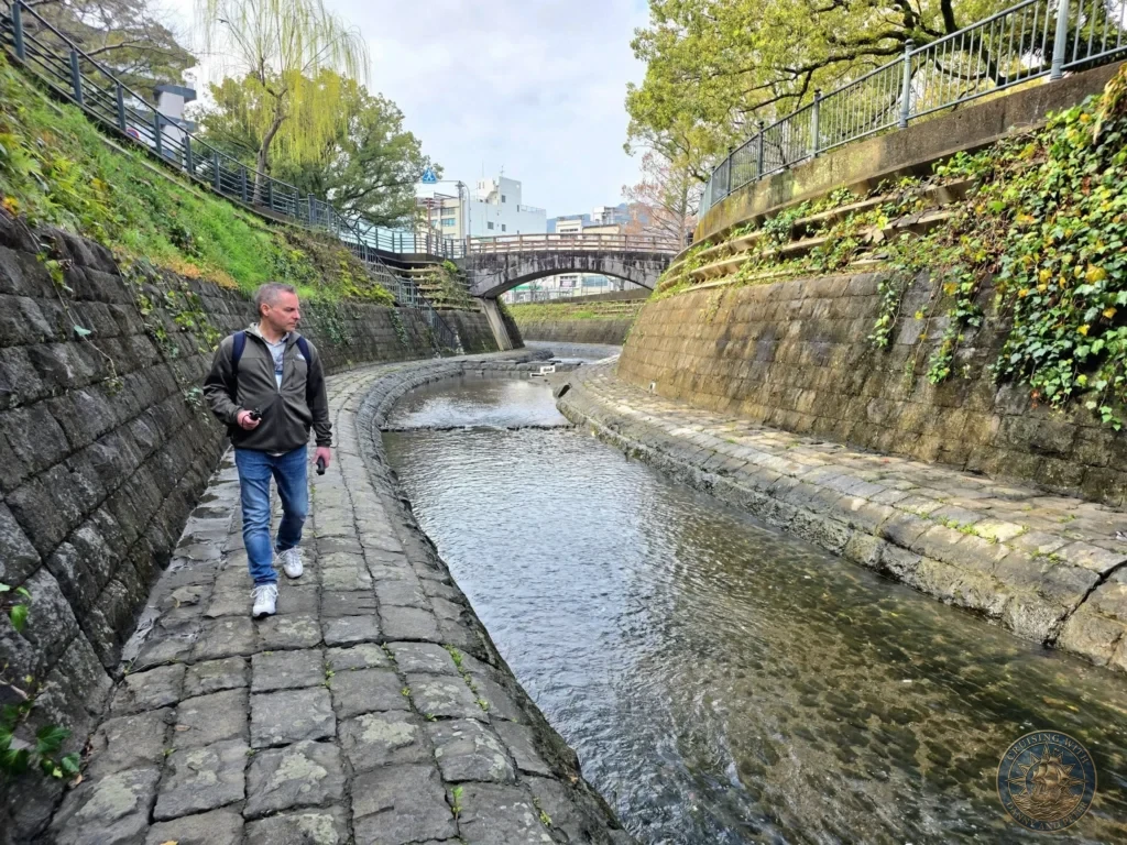 Peter walking along the stone path beside a historic canal near Hypocenter Park in Nagasaki, Japan. Stone retaining walls, bridges, and greenery line the waterway in this peaceful area, featured in Nagasaki Japan cruise port guide, things to do in Nagasaki from cruise ship, and one day in Nagasaki Japan itinerary from the Westerdam. by Danny and Peter.