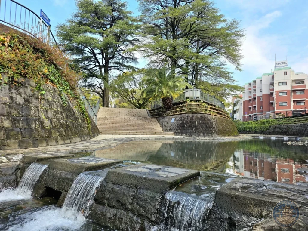 Peaceful canal with small waterfalls and stone embankments at Hypocenter Park in Nagasaki, Japan, surrounded by trees, stairs, and city buildings including the Seagull hotel in the background. Featured in Nagasaki Japan cruise port guide, things to do in Nagasaki from cruise ship, and one day in Nagasaki Japan itinerary from the Westerdam. by Danny and Peter.