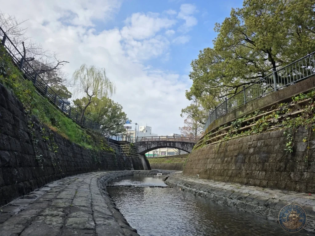 Scenic view of the stone-lined canal at Hypocenter Park in Nagasaki, Japan, with cobblestone paths, high stone retaining walls covered in ivy, trees, and a bridge in the distance under a blue sky. Featured in Nagasaki Japan cruise port guide, things to do in Nagasaki from cruise ship, and one day in Nagasaki Japan itinerary from the Westerdam. by Danny and Peter.