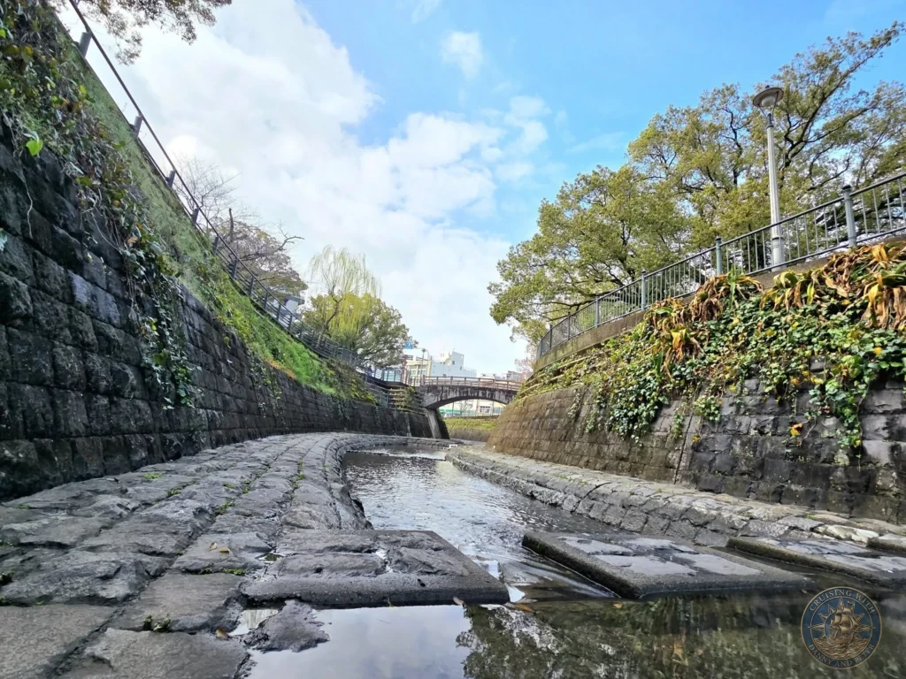 Stone-paved path and canal with reflective water at Hypocenter Park in Nagasaki, Japan, flanked by high stone walls covered in greenery and trees under a bright blue sky. A quiet, reflective area near the atomic bombing site. Featured in Nagasaki Japan cruise port guide, things to do in Nagasaki from cruise ship, and one day in Nagasaki Japan itinerary from the Westerdam. by Danny and Peter.