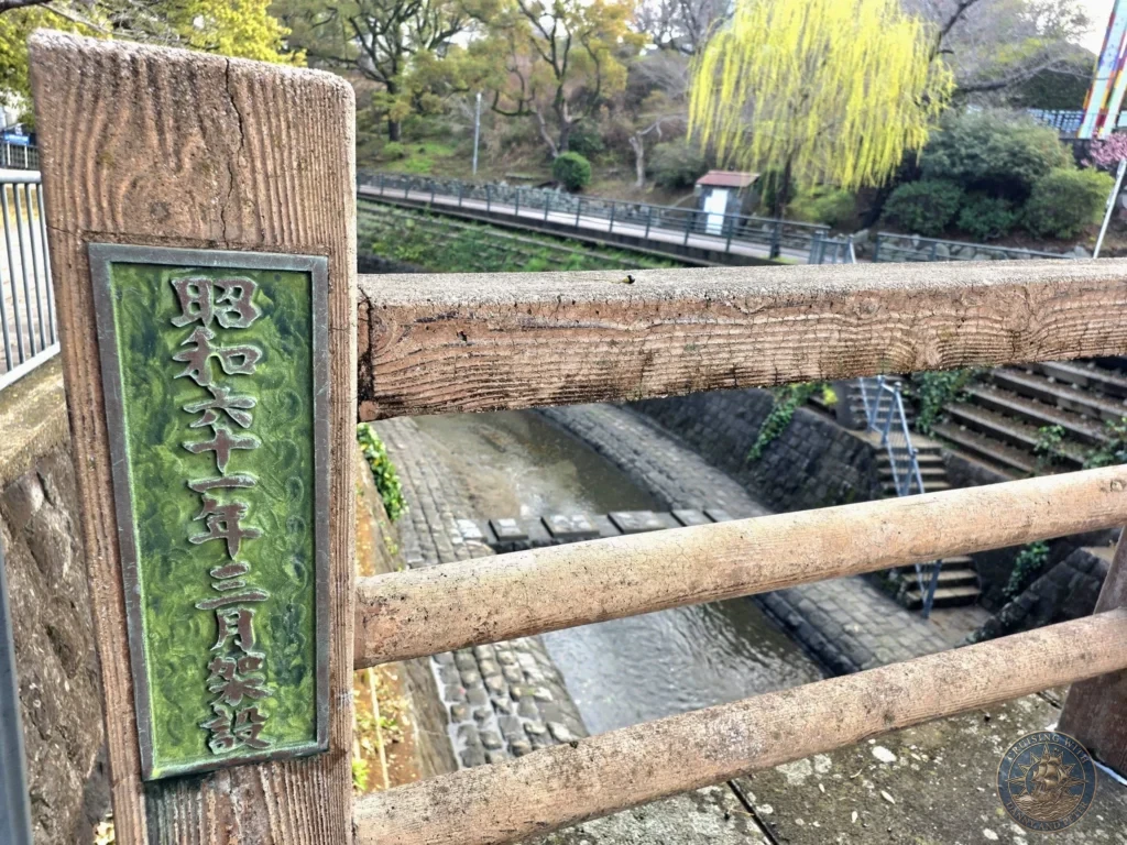 Close-up of a weathered wooden bridge railing with a green bronze plaque bearing Japanese text at Hypocenter Park in Nagasaki, Japan. The background shows the historic stone canal, trees, and pathways. Featured in Nagasaki Japan cruise port guide, things to do in Nagasaki from cruise ship, and one day in Nagasaki Japan itinerary from the Westerdam. by Danny and Peter.