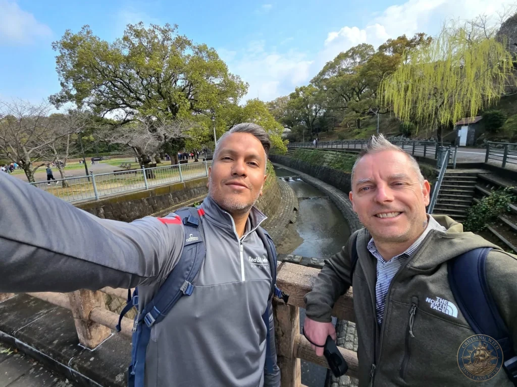 Selfie of Danny and Peter on a wooden bridge at Hypocenter Park in Nagasaki, Japan, with the peaceful stone-lined canal, lush green trees, and park pathways in the background. A reflective stop near the atomic bombing epicenter. Featured in Nagasaki Japan cruise port guide, things to do in Nagasaki from cruise ship, and one day in Nagasaki Japan itinerary from the Westerdam. by Danny and Peter.