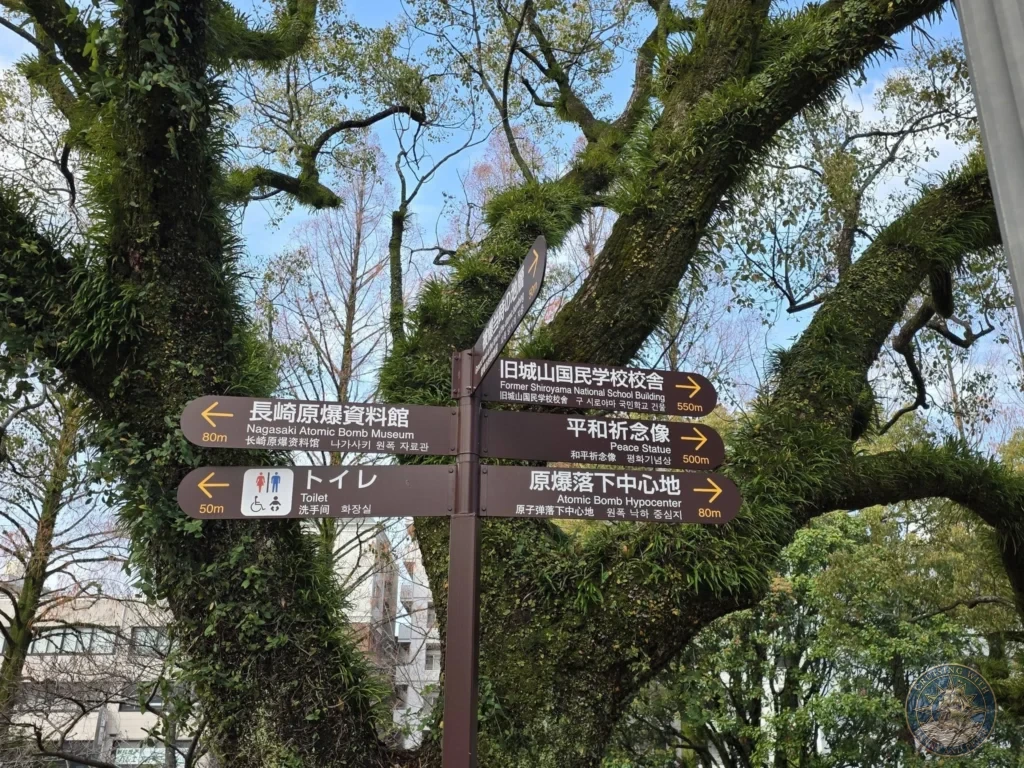 Directional signpost at Hypocenter Park in Nagasaki, Japan, showing distances to the Nagasaki Atomic Bomb Museum, Atomic Bomb Hypocenter, Peace Statue, Former Shiroyama National School Building, and toilets. The sign is framed by large, moss-covered trees under a blue sky. Featured in Nagasaki Japan cruise port guide, things to do in Nagasaki from cruise ship, and one day in Nagasaki Japan itinerary from the Westerdam. by Danny and Peter.