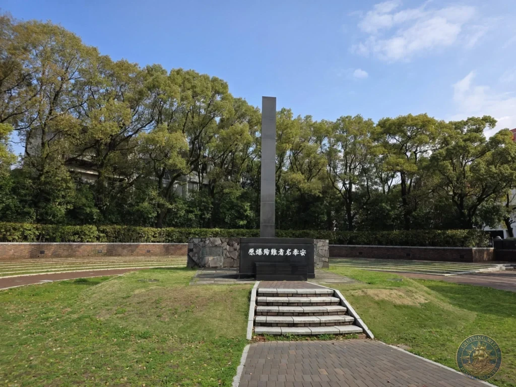 Tall stone monument dedicated to the atomic bomb victims at Hypocenter Park in Nagasaki, Japan, set on a grassy hill with stone steps and surrounded by lush green trees under a clear blue sky. Featured in Nagasaki Japan cruise port guide, things to do in Nagasaki from cruise ship, and one day in Nagasaki Japan itinerary from the Westerdam. by Danny and Peter.