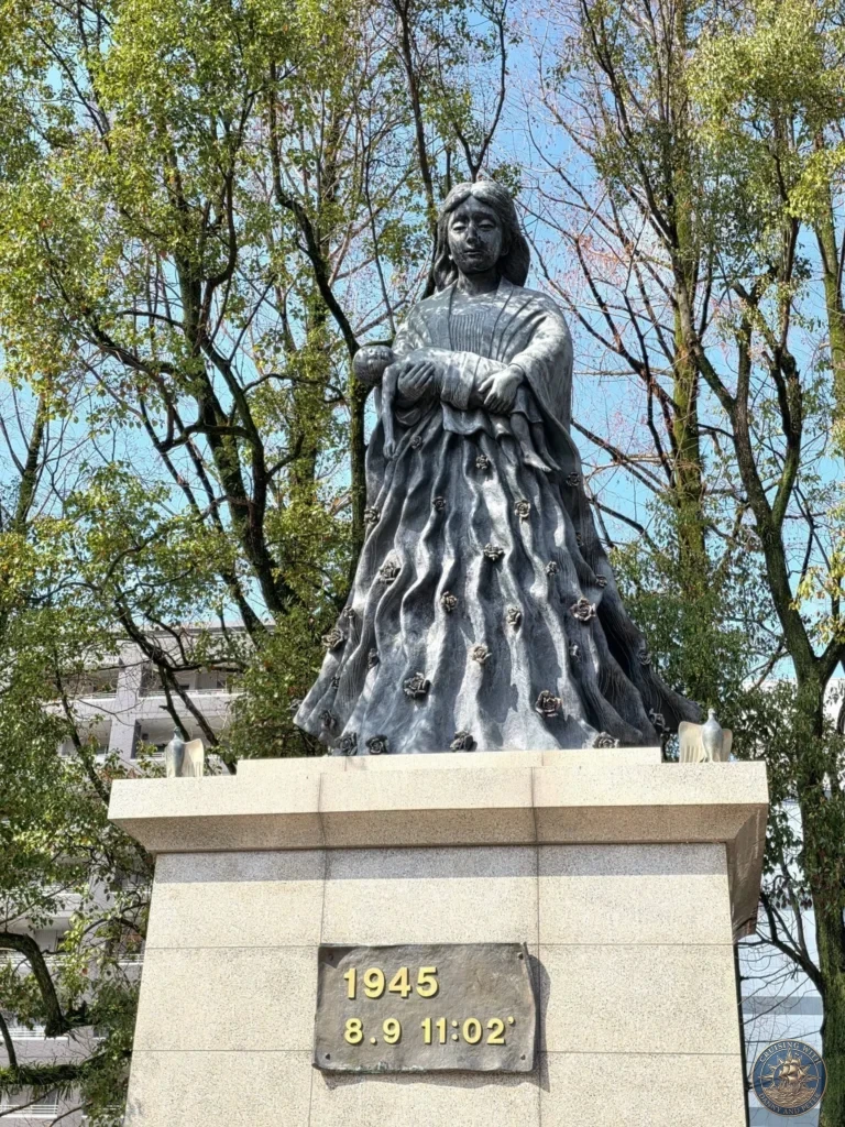 Bronze statue of a mother cradling her child at Hypocenter Park in Nagasaki, Japan, with the exact time of the atomic bombing (1945.8.9 11:02) inscribed on the base. Surrounded by trees under a clear blue sky. Featured in Nagasaki Japan cruise port guide, things to do in Nagasaki from cruise ship, and one day in Nagasaki Japan itinerary from the Westerdam. by Danny and Peter.