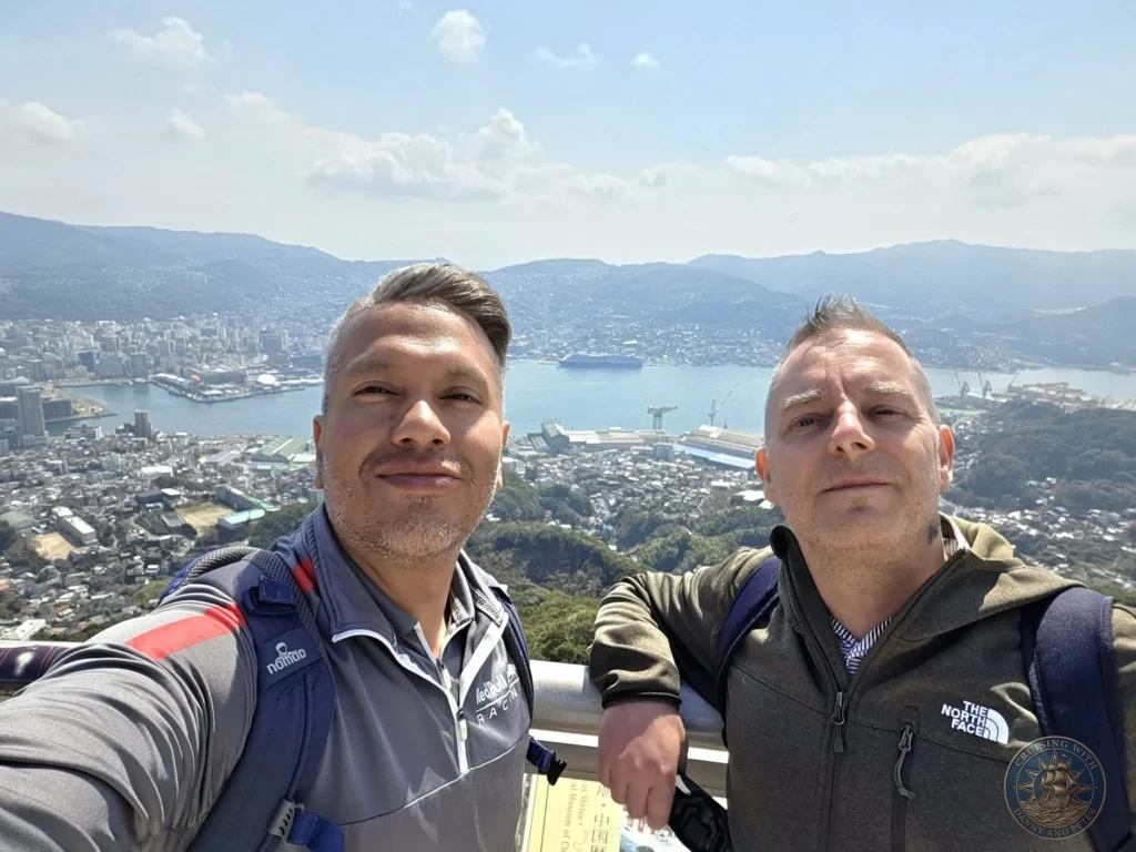 Selfie of Danny and Peter at the Mount Inasa observation deck in Nagasaki, Japan, with a stunning panoramic view of the city, harbor, and a large cruise ship, the Westerdam, docked below. Featured in Nagasaki Japan cruise port guide, things to do in Nagasaki from cruise ship, and one day in Nagasaki Japan itinerary from the Westerdam. by Danny and Peter.