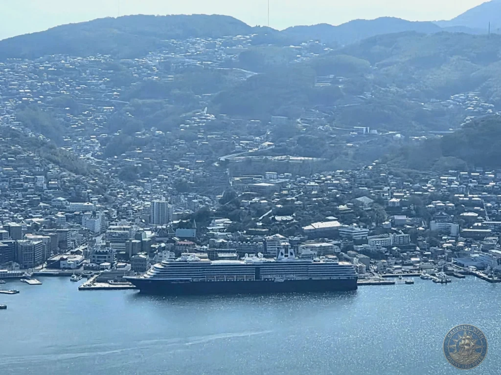 Panoramic view from Mount Inasa overlooking Nagasaki harbor with the MS Westerdam cruise ship docked prominently in the foreground. The city sprawls across the hills and coastline with mountains in the background. Featured in Nagasaki Japan cruise port guide, things to do in Nagasaki from cruise ship, and one day in Nagasaki Japan itinerary from the Westerdam. by Danny and Peter.