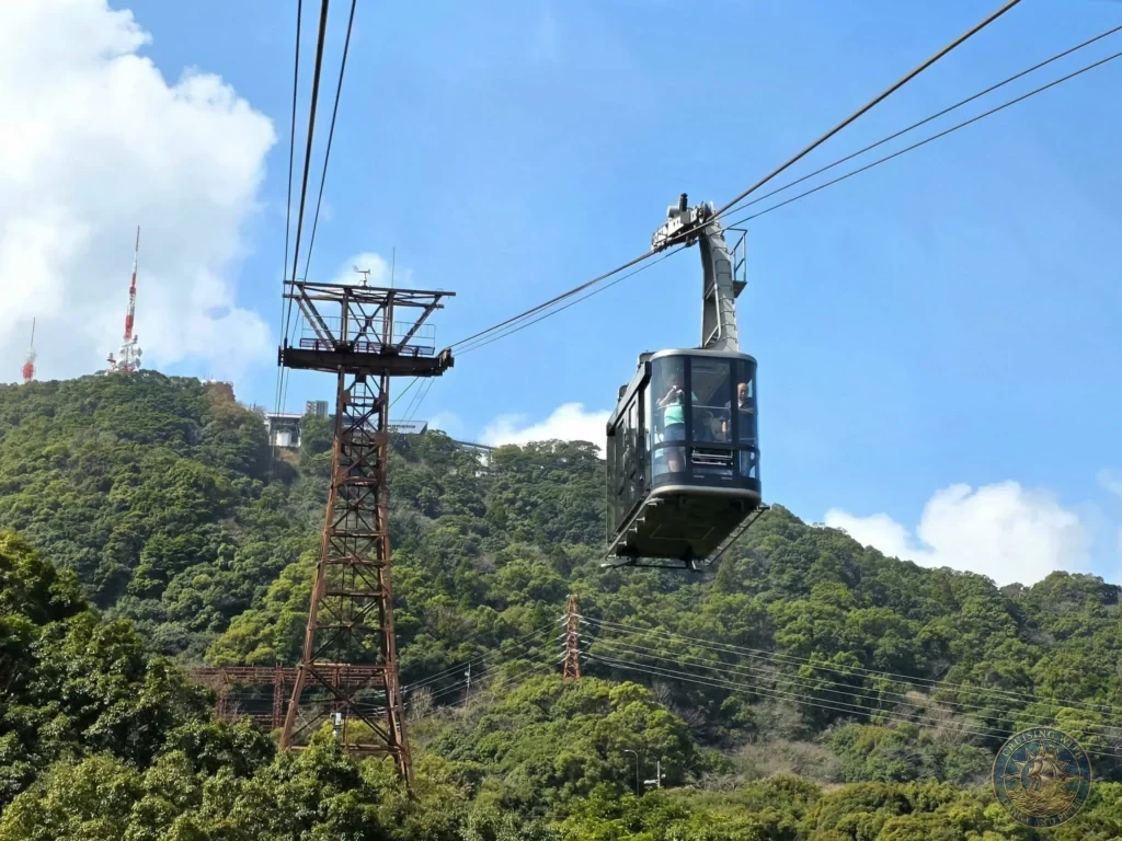 Ropeway gondola carrying passengers up Mount Inasa in Nagasaki, Japan, with the steel support tower and lush green hillside in the background under a blue sky. Featured in Nagasaki Japan cruise port guide, things to do in Nagasaki from cruise ship, and one day in Nagasaki Japan itinerary from the Westerdam. by Danny and Peter.