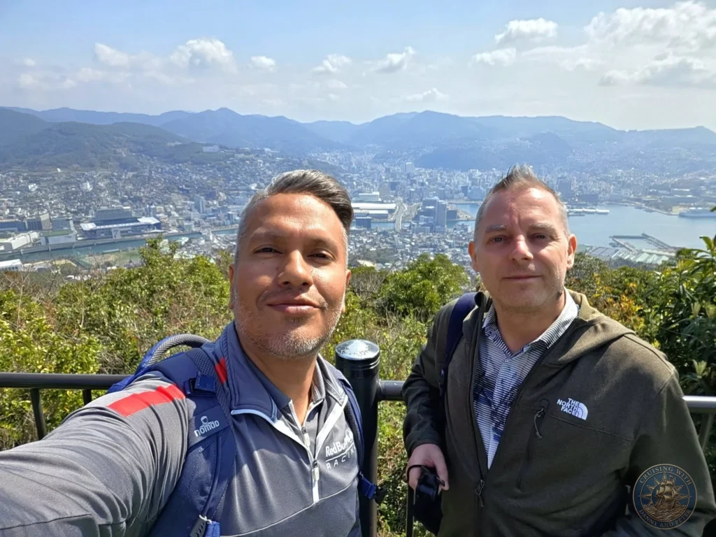Selfie of Danny and Peter at the Mount Inasa observation deck in Nagasaki, Japan, with a sweeping panoramic view of the city, harbor, and surrounding mountains behind them. Featured in Nagasaki Japan cruise port guide, things to do in Nagasaki from cruise ship, and one day in Nagasaki Japan itinerary from the Westerdam. by Danny and Peter.