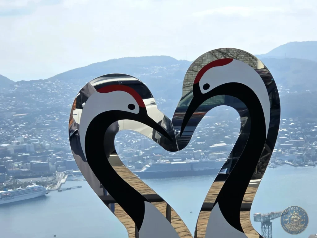 Modern heart-shaped sculpture made of two mirrored cranes at the Mount Inasa observation deck in Nagasaki, Japan, framing a panoramic view of the city, harbor, and cruise ships below. Featured in Nagasaki Japan cruise port guide, things to do in Nagasaki from cruise ship, and one day in Nagasaki Japan itinerary from the Westerdam. by Danny and Peter.