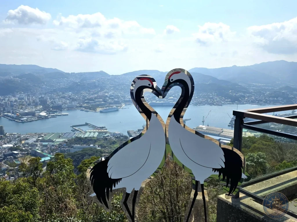 Large heart-shaped sculpture of two cranes at the Mount Inasa viewpoint in Nagasaki, Japan, perfectly framing the panoramic view of the city, harbor, and cruise ships below. Featured in Nagasaki Japan cruise port guide, things to do in Nagasaki from cruise ship, and one day in Nagasaki Japan itinerary from the Westerdam. by Danny and Peter.
