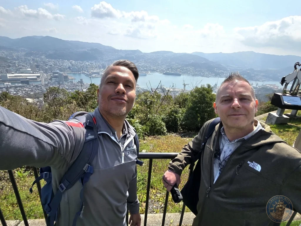 Selfie of Danny and Peter at the Mount Inasa viewpoint in Nagasaki, Japan, with a stunning panoramic backdrop of the city, harbor, and distant mountains under a bright blue sky. Featured in Nagasaki Japan cruise port guide, things to do in Nagasaki from cruise ship, and one day in Nagasaki Japan itinerary from the Westerdam. by Danny and Peter.