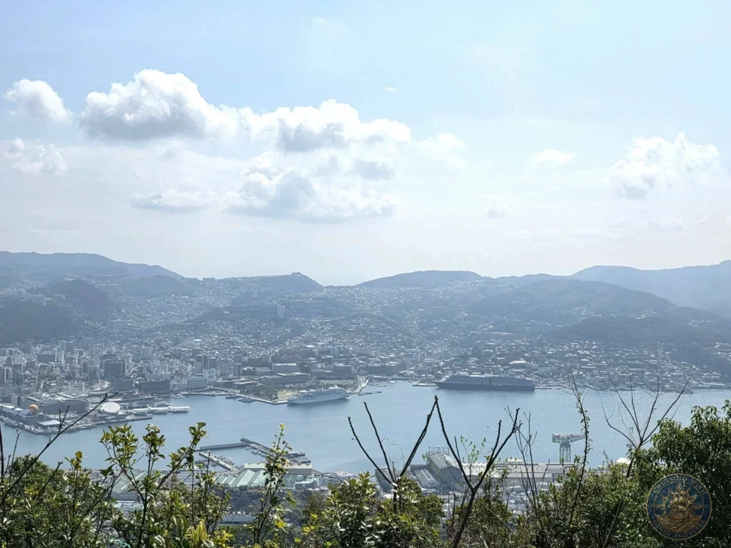 Wide panoramic view from Mount Inasa overlooking Nagasaki city, harbor, and two cruise ships docked below, with green hills and mountains in the background under a bright blue sky with scattered clouds. Featured in Nagasaki Japan cruise port guide, things to do in Nagasaki from cruise ship, and one day in Nagasaki Japan itinerary from the Westerdam. by Danny and Peter.