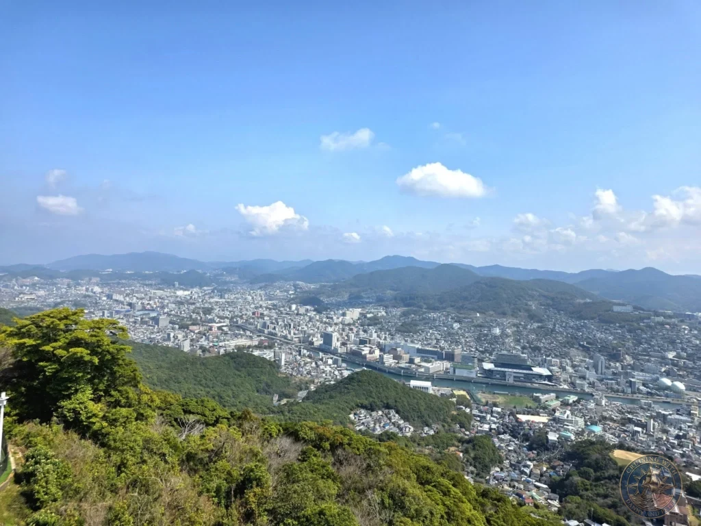Panoramic view from Mount Inasa overlooking the sprawling city of Nagasaki, Japan, with green forested hills in the foreground and distant mountains under a bright blue sky. Featured in Nagasaki Japan cruise port guide, things to do in Nagasaki from cruise ship, and one day in Nagasaki Japan itinerary from the Westerdam. by Danny and Peter.