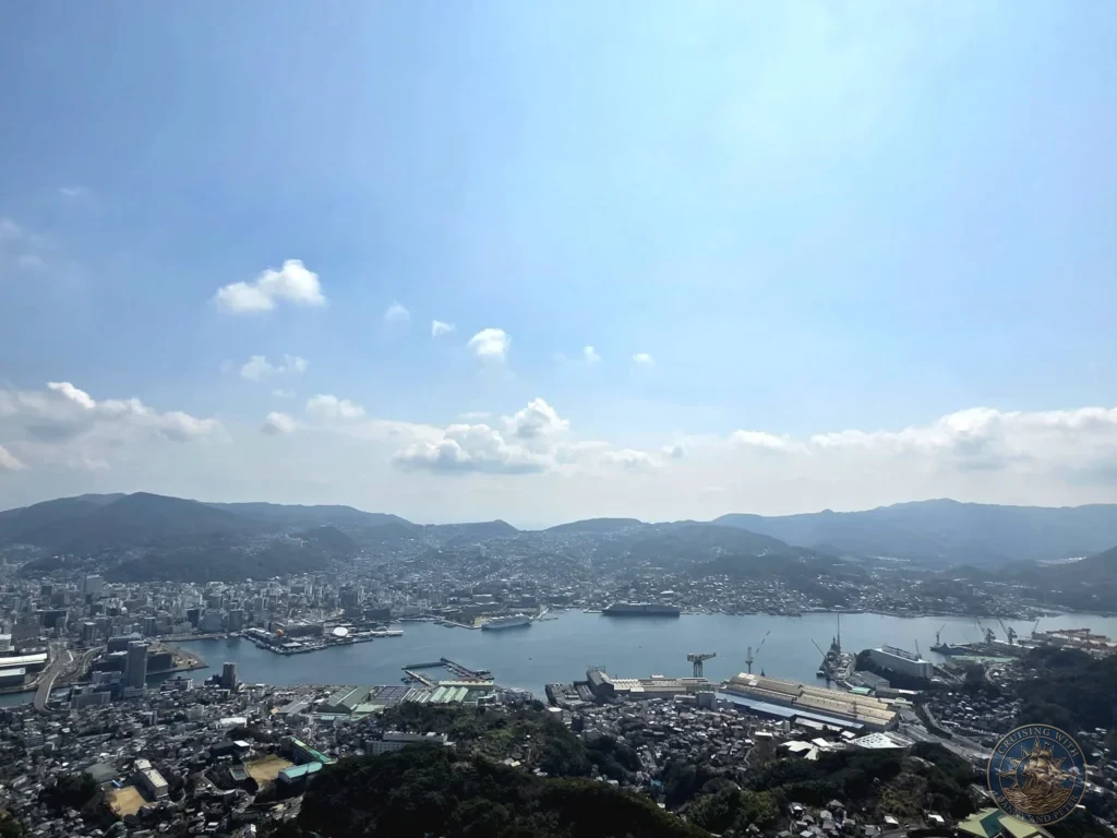 Expansive panoramic view from Mount Inasa showing Nagasaki city, the wide harbor with two large cruise ships docked, industrial port facilities, cranes, and surrounding green mountains under a bright blue sky. Featured in Nagasaki Japan cruise port guide, things to do in Nagasaki from cruise ship, and one day in Nagasaki Japan itinerary from the Westerdam. by Danny and Peter.
