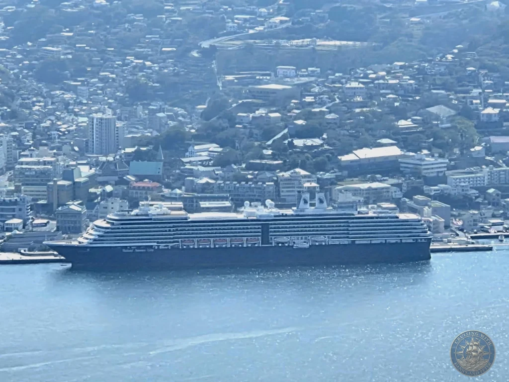 Aerial view from Mount Inasa of the Holland America Line Westerdam cruise ship docked in Nagasaki harbor, Japan, with the city and green hills in the background. Featured in Nagasaki Japan cruise port guide, things to do in Nagasaki from cruise ship, and one day in Nagasaki Japan itinerary from the Westerdam. by Danny and Peter.