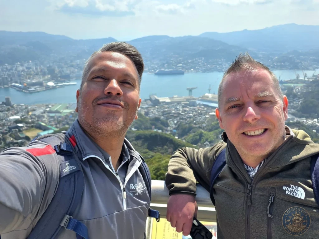 Selfie of Danny and Peter smiling at the Mount Inasa observation deck in Nagasaki, Japan, with a spectacular panoramic view of the city, harbor, and distant mountains behind them under a bright blue sky. Featured in Nagasaki Japan cruise port guide, things to do in Nagasaki from cruise ship, and one day in Nagasaki Japan itinerary from the Westerdam. by Danny and Peter.