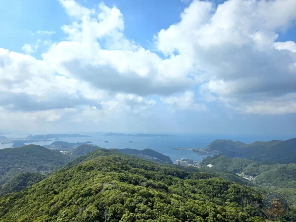 Panoramic view from Mount Inasa showing lush green forested hills in the foreground, the city of Nagasaki, and the wide blue sea with distant islands on the horizon under a bright blue sky with scattered clouds. Featured in Nagasaki Japan cruise port guide, things to do in Nagasaki from cruise ship, and one day in Nagasaki Japan itinerary from the Westerdam. by Danny and Peter.